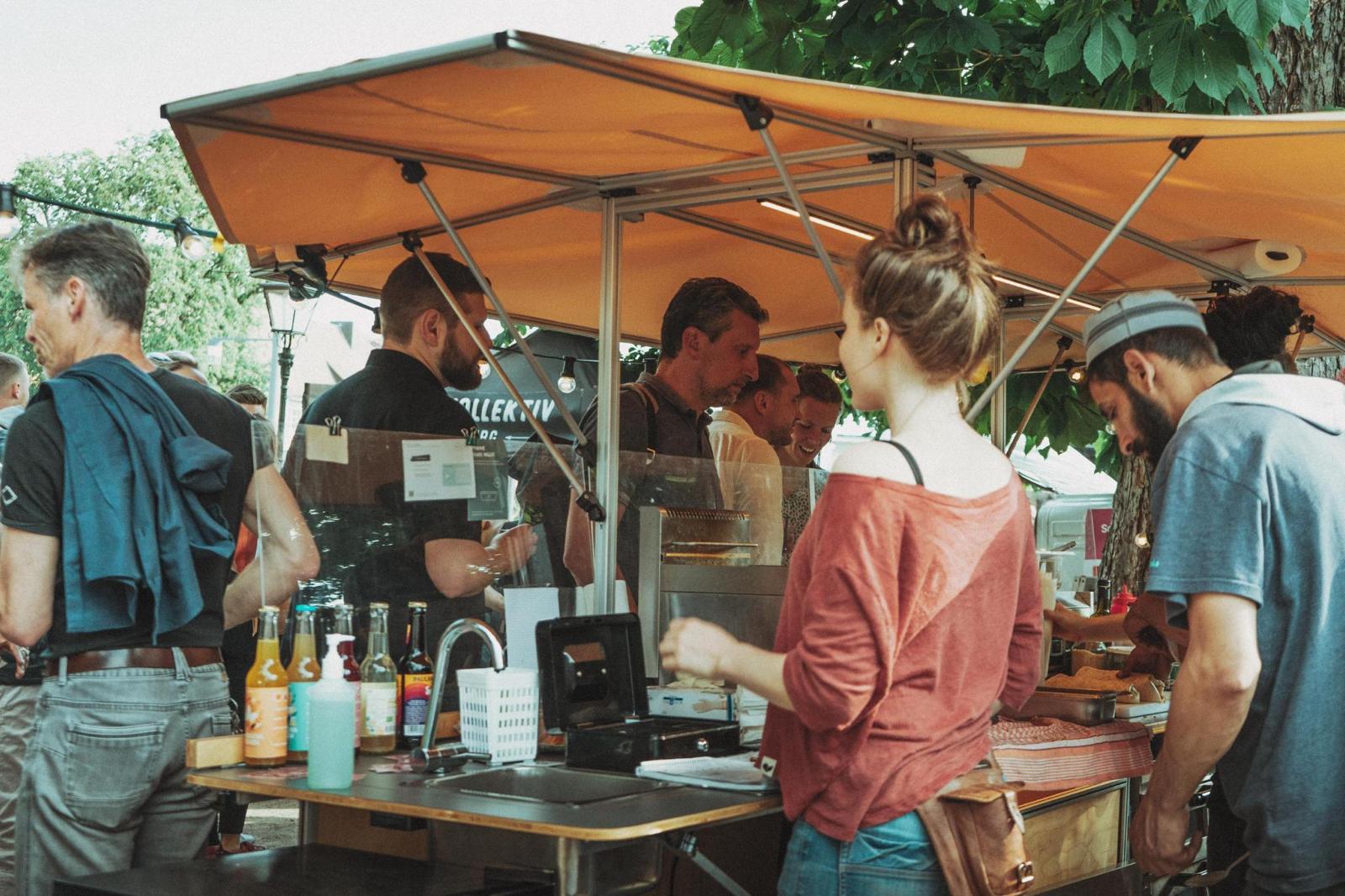Belebter Marktstand unter orangefarbenem Sonnenschirm: Gäste und Betreiber einer mobilen Gastronomie im Gespräch