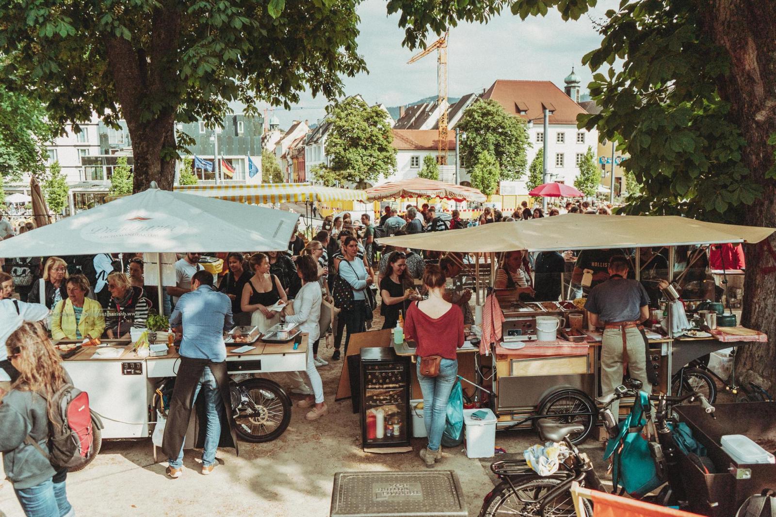 Zwei mobile CARLA-Verkaufsstände auf einem belebten Wochenmarkt zwischen Bäumen und historischen Gebäuden